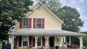 A two-story house with a porch, numbered 100, surrounded by greenery and potted plants.