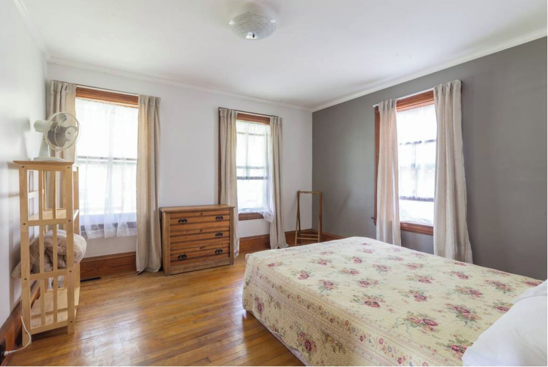 Ein Schlafzimmer mit einem hölzernen Schminktisch, einem Bett mit floralem Überwurf und zwei Fenstern mit Vorhängen.