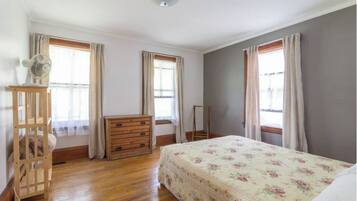 Ein Schlafzimmer mit einem hölzernen Schminktisch, einem Bett mit floralem Überwurf und zwei Fenstern mit Vorhängen.