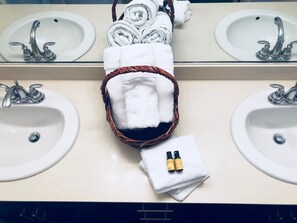 A bathroom with two sinks, a mirror, and a basket containing rolled towels and a small towel with two bottles.
