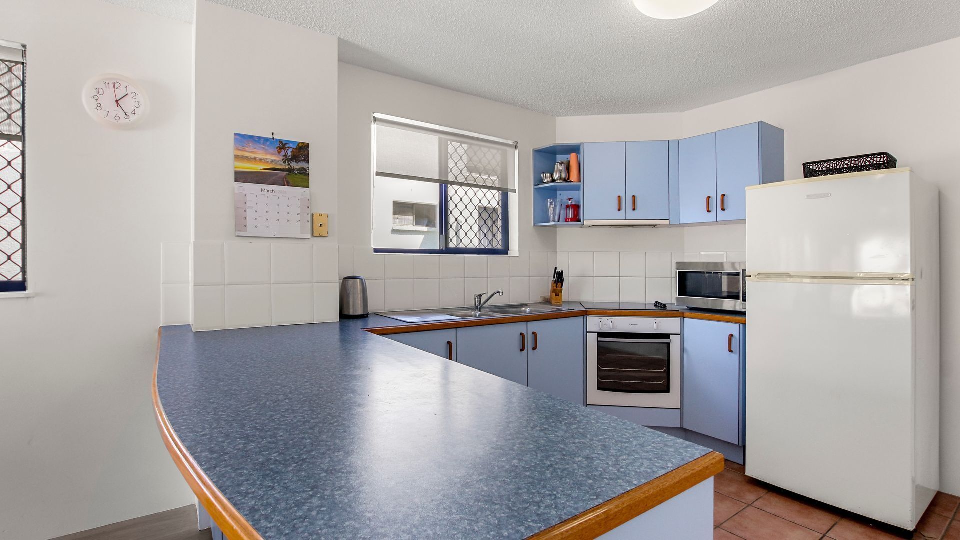 Una cocina con encimera azul, armarios blancos, un refrigerador, un microondas y una ventana con vistas.