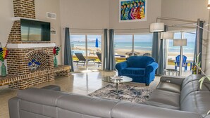 Uma sala de estar ampla com lareira, um sofá grande, uma poltrona azul e vista para a praia.