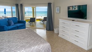 Quarto de hotel com uma cama grande, um sofá azul, uma TV e vista para a praia.
