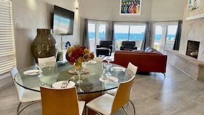 A dining area with a glass table, chairs, a vase with flowers, a flat-screen TV, and a view of the sea through large windows. - Villa 5 – Newly Remodeled Oceanfront Luxury Villa (Puerto Peñasco)