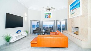A modern living room with a large orange sofa, a flat-screen TV, a ceiling fan, and a fireplace. - Villa 5 – Newly Remodeled Oceanfront Luxury Villa (Puerto Peñasco)