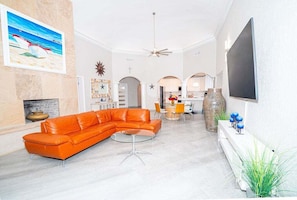 Uma sala de estar com sofá seccional, televisão, lareira e cozinha ao fundo.