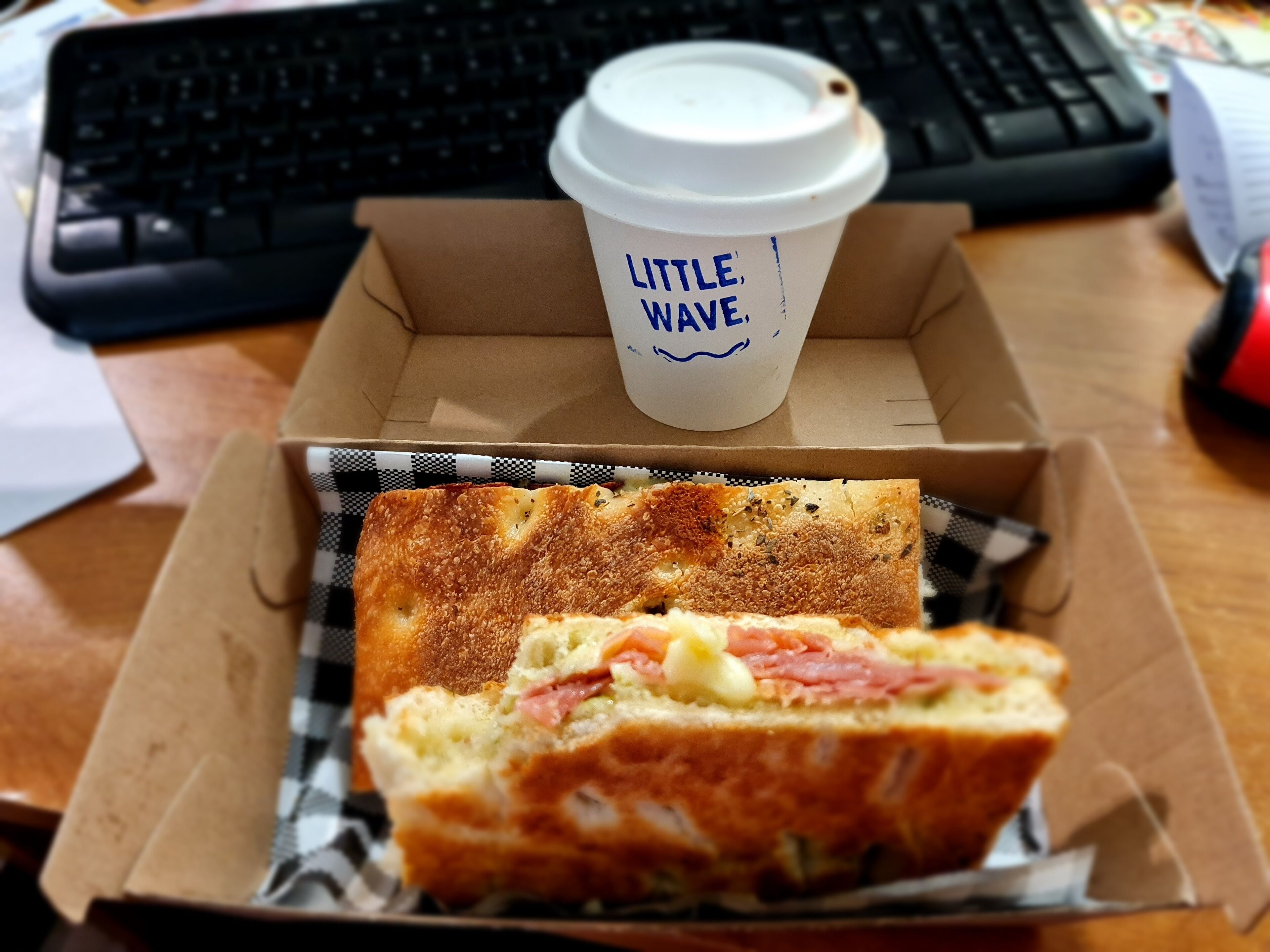 A sandwich in a cardboard box with a coffee cup labeled "Little Wave" on a desk.