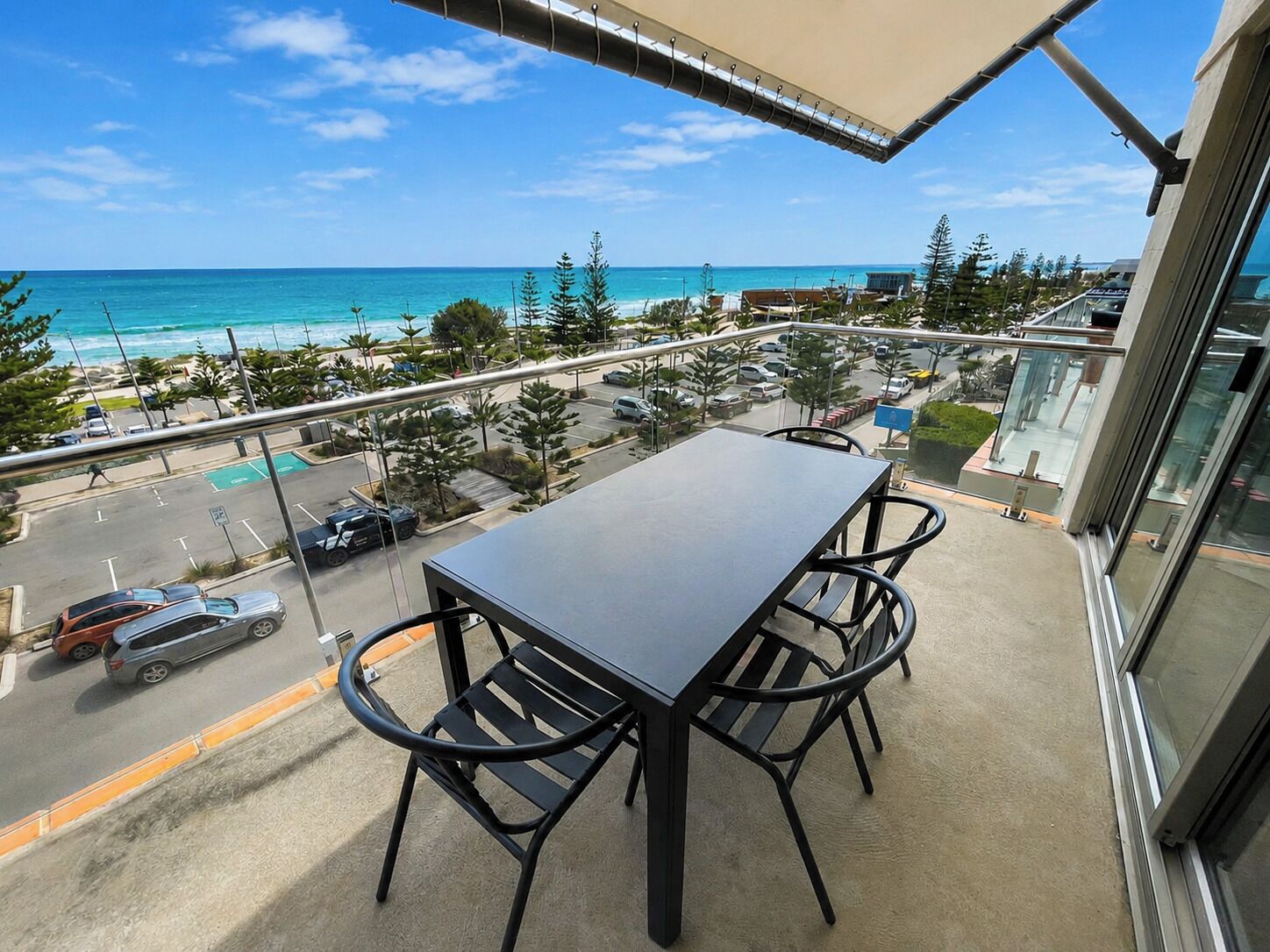 A balcony with a table and chairs, overlooking a parking area and a view of the ocean.