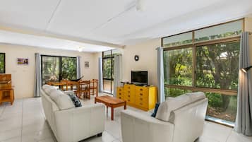 A spacious living room with two white sofas, a wooden dining table, and a yellow dresser. Large windows offer a view of greenery outside.