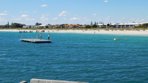 A beach with a pier extending into the water, surrounded by houses and a clear sky.
