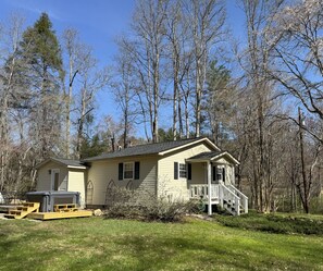 Une maison de plain-pied avec un porche à l’avant, entourée d’arbres et d’une pelouse.