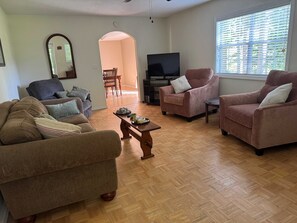 Una sala de estar con un sofá, dos sillones, una mesa de centro, un televisor y una ventana con persianas.