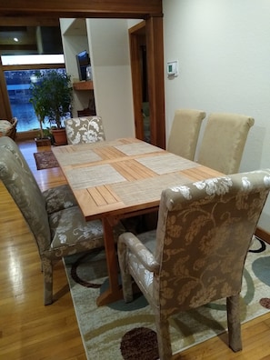 A dining area with a wooden table and patterned chairs, a potted plant, and a view of a body of water through a window.