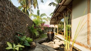 A stone pathway leading to a thatched-roof structure, surrounded by tropical plants and a decorative water feature.