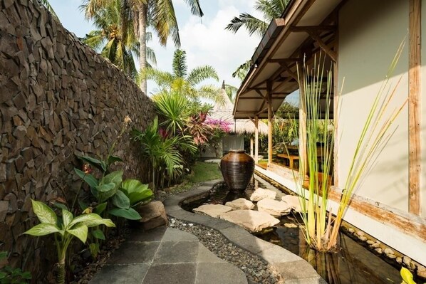 Un sendero de piedra conduce a una construcción con techo de paja, rodeada de plantas tropicales y una fuente decorativa.