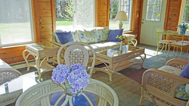Una habitación acogedora con muebles de mimbre, una mesa de centro de vidrio con flores y ventanas amplias que ofrecen vistas al exterior.