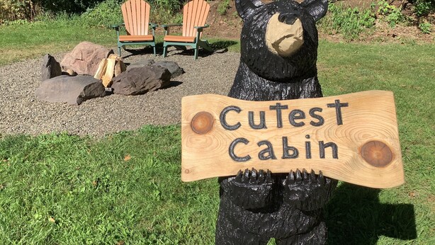 Ein Bär hält ein Schild mit der Aufschrift „Cutest Cabin“ in einem Garten mit zwei Stühlen und Steinen.