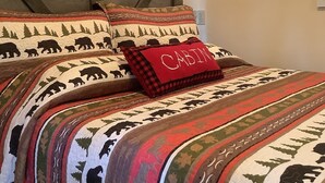 Ein Schlafzimmer mit einem Bett, ausgestattet mit einer Bären-Quiltdecke, einem Kissen mit der Aufschrift 'Cabin' und einem Schild über dem Bett mit der Aufschrift 'Möge Frieden mit dir sein, während du hier bist'.