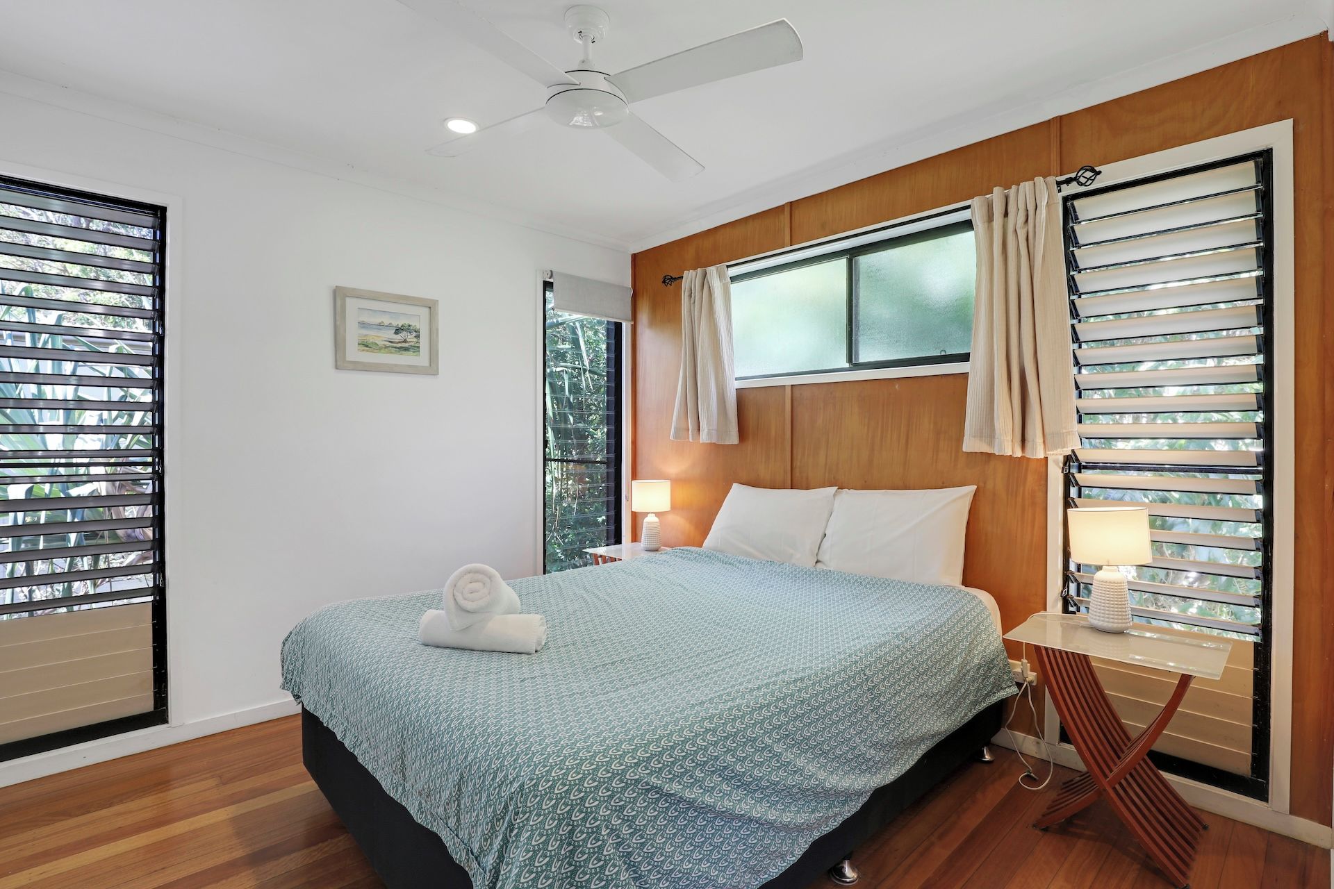 A bedroom with a bed, a ceiling fan, a window with curtains, and a bedside table with a lamp.