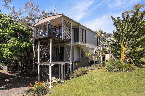 Une maison à deux étages avec un balcon à l’étage supérieur, entourée de verdure et d’une pelouse bien entretenue.