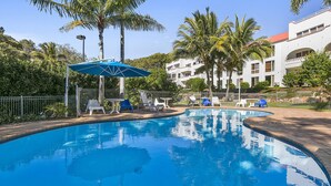 Une piscine entourée de chaises longues et de parasols, bordée de palmiers et d’un bâtiment blanc doté de balcons.