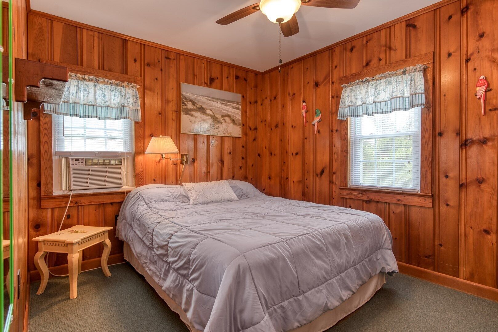 A bedroom with wooden paneling, a bed with a grey duvet, two windows with lace curtains, a ceiling fan, and a small bedside table.