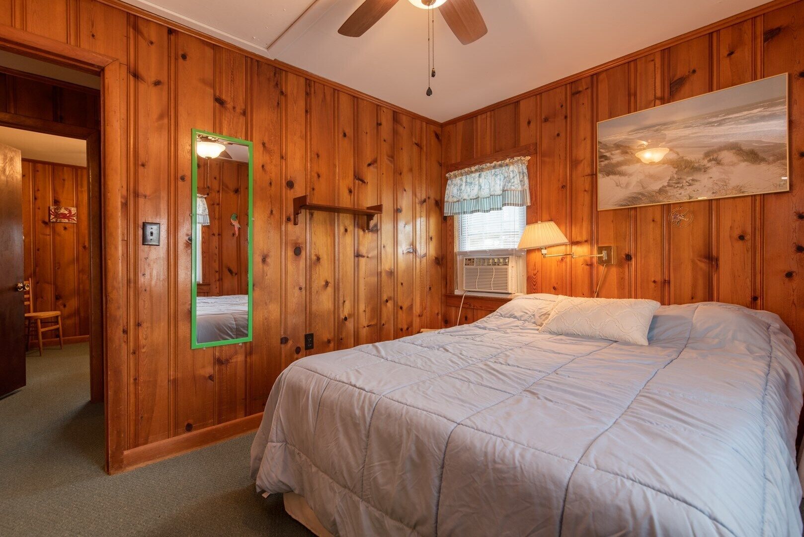 A bedroom with wooden paneling, a bed with white bedding, a mirror with a green frame, a window with a lace curtain, and a painting on the wall.