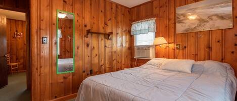 A bedroom with wooden paneling, a bed with white bedding, a mirror with a green frame, a window with a lace curtain, and a painting on the wall.