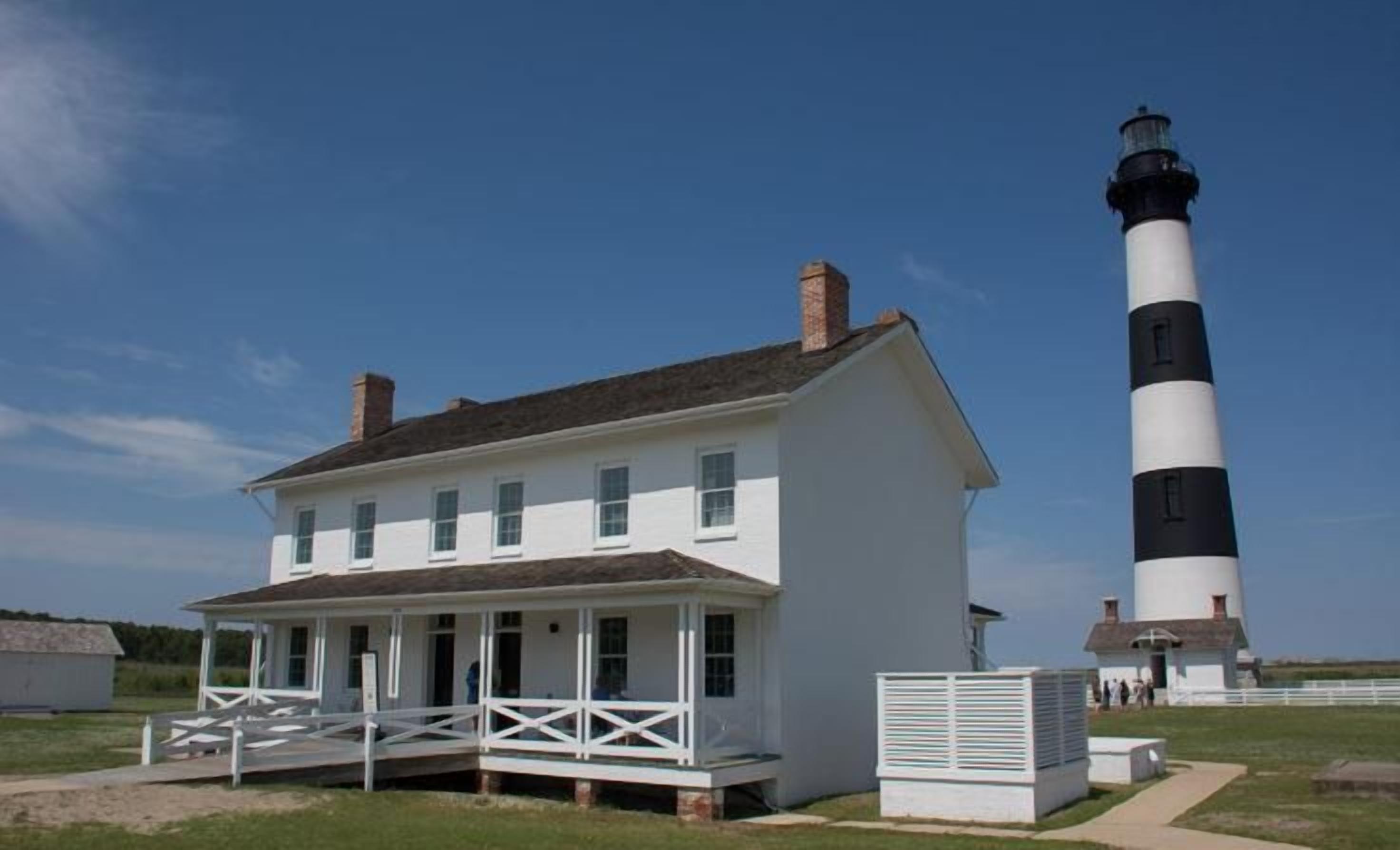 Une maison blanche de style colonial, dotée d’un porche, et un phare noir et blanc en arrière-plan.