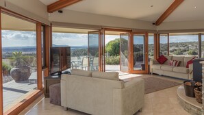 A spacious living room with large sliding glass doors leading to an outdoor patio, a beige sofa, a television, and a fireplace. - Almonta Park Lodge (Coffin Bay)