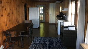 Un interior de madera con una mesa y sillas para comer, una zona de cocina con estufa y fregadero, y una puerta que da al exterior.