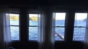 Habitación con tres ventanas que dan a una masa de agua, una terraza de madera afuera y una silla azul adentro.