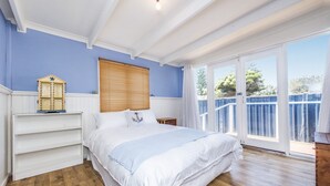 A bedroom with a large bed, a wooden floor, a window with blinds, and a balcony with a metal railing. - Doc's Cottage Lancelin - Ocean Views - Pet Friendly. (Lancelin)