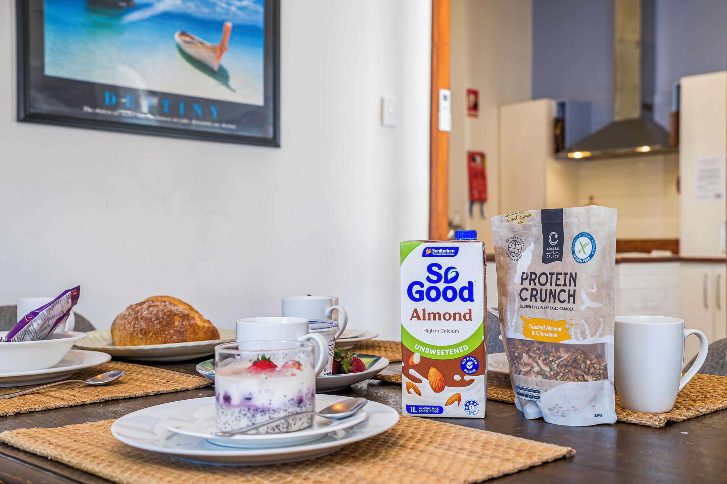 A breakfast setup with a bowl of cereal, a glass of yogurt with fruit, a loaf of bread, and two protein bars on a table.