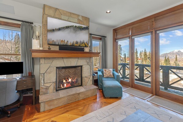 A living room with a stone fireplace, a television mounted above it, and a view of a snowy landscape through large windows.