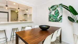 Un'area pranzo moderna con un tavolo di legno, sedie bianche, una ciotola nera e un grande dipinto con una foglia tropicale sulla parete.