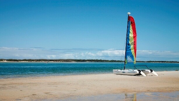 Un voilier aux voiles colorées sur une plage de sable, avec une eau calme et un ciel dégagé.