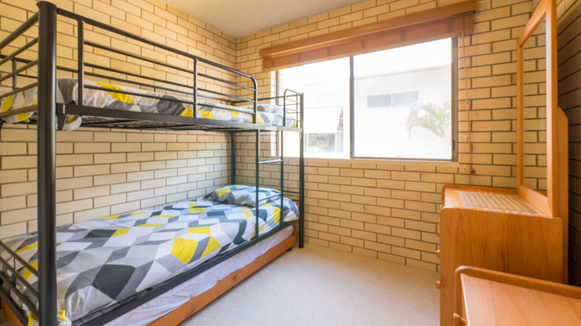 Habitación tipo dormitorio con literas, un escritorio de madera y una ventana con vistas a un edificio.