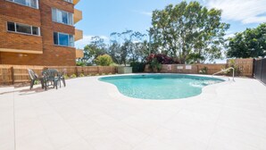 Une piscine avec une terrasse carrelée, entourée d’une clôture en bois, et un bâtiment en briques en arrière-plan.