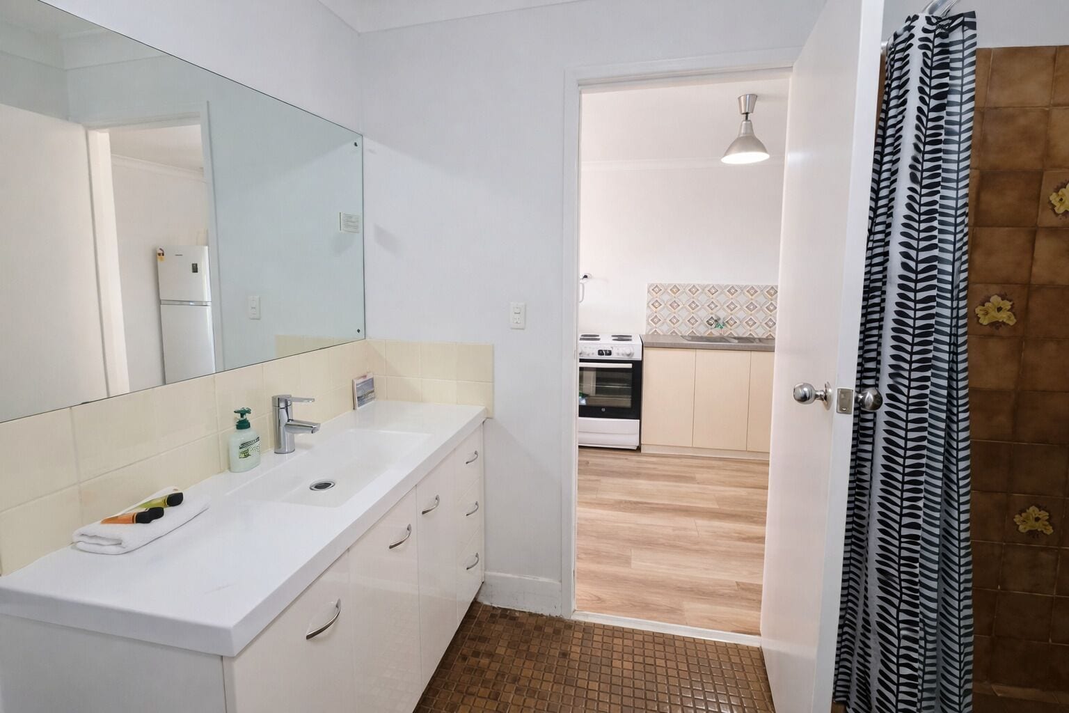 A bathroom with a white sink, mirror, and cabinets. There is a door leading to a kitchen with a stove and tiled backsplash.