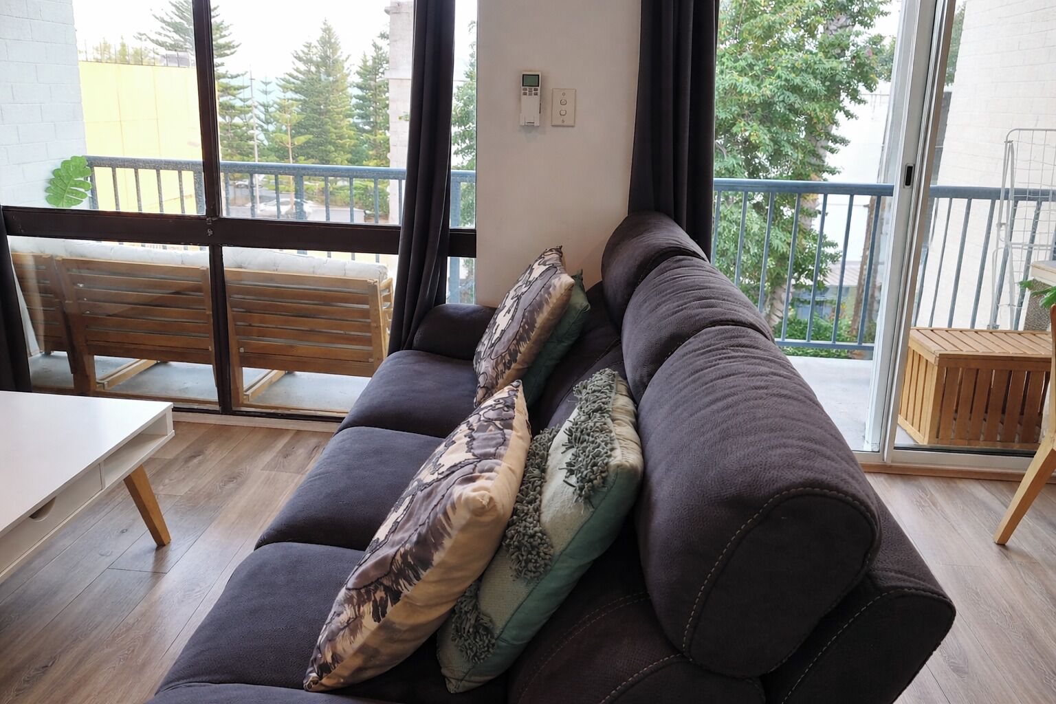A modern living room with a dark sofa, patterned cushions, a wooden bench, and a view through large windows.