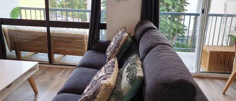 A modern living room with a dark sofa, patterned cushions, a wooden bench, and a view through large windows.
