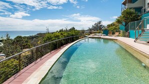 A swimming pool with a curved edge, surrounded by a metal railing, and overlooking a scenic view of the ocean and hills. - Panoramic ocean and beach views from this beautifully appointed two... (Point Lookout)
