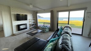 A modern living room with a fireplace, a flat-screen TV, a sofa, and a large window offering a view of the sea. - Sandy Shores on Irby - Beach frontage at beautiful Sisters Beach (Sisters Beach)