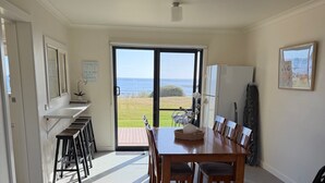 A dining area with a table and chairs, a view of the sea through a sliding door, a small plant on a shelf, and a refrigerator. - Sandy Shores on Irby - Beach frontage at beautiful Sisters Beach (Sisters Beach)