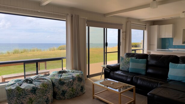 A living room with a view of the ocean, a black leather sofa, and a glass coffee table. - Sandy Shores on Irby - Beach frontage at beautiful Sisters Beach (Sisters Beach)