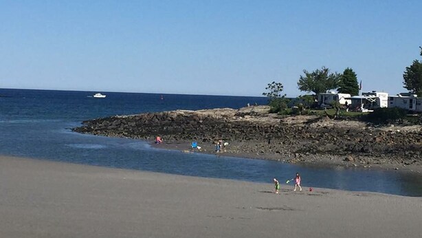 Ein Strand mit felsiger Küste, einigen Personen und einem weißen Boot im Wasser. Auf dem Strand stehen Wohnmobile geparkt.