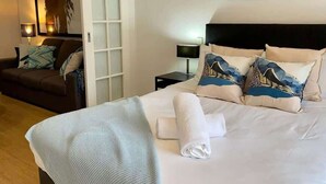 Ein ordentlich bezogenes Bett mit weißer Decke und gerollten Handtüchern, ein dunkelbraunes Sofa mit gemusterten Kissen, ein kleiner Nachttisch mit Lampe sowie ein gerahmtes Kunstwerk an der Wand.