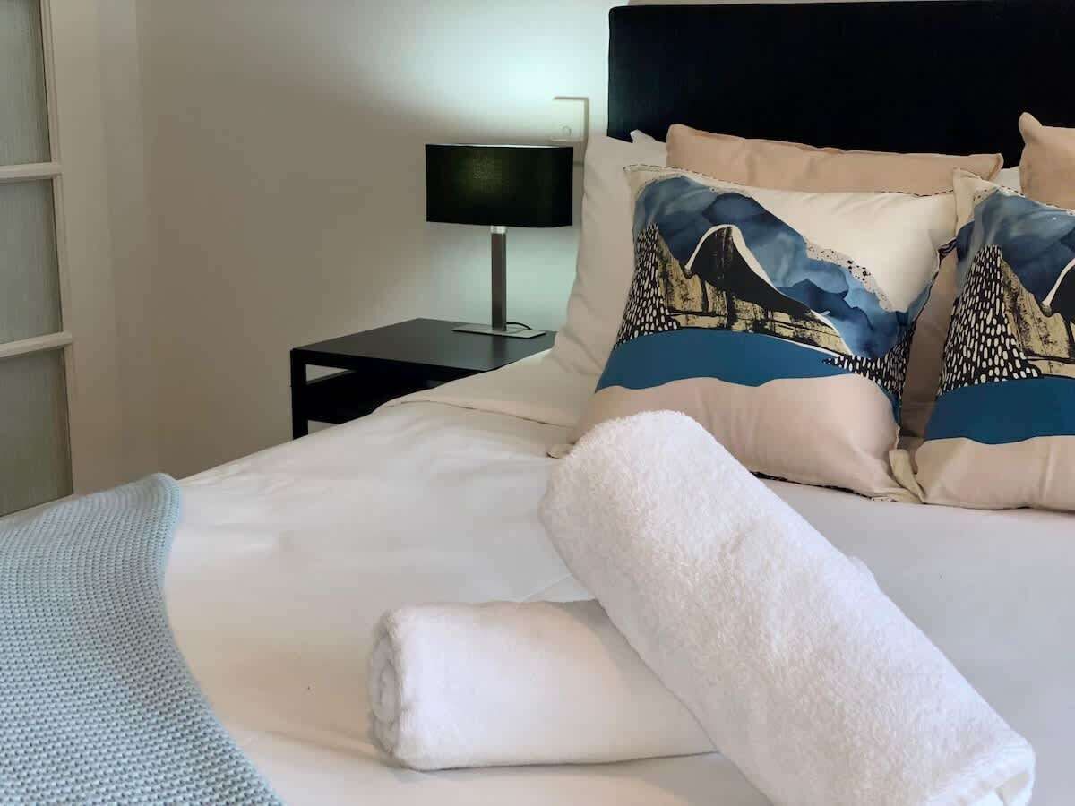 A neatly made bed with white linens, a rolled white towel, and decorative pillows with a blue and beige pattern.