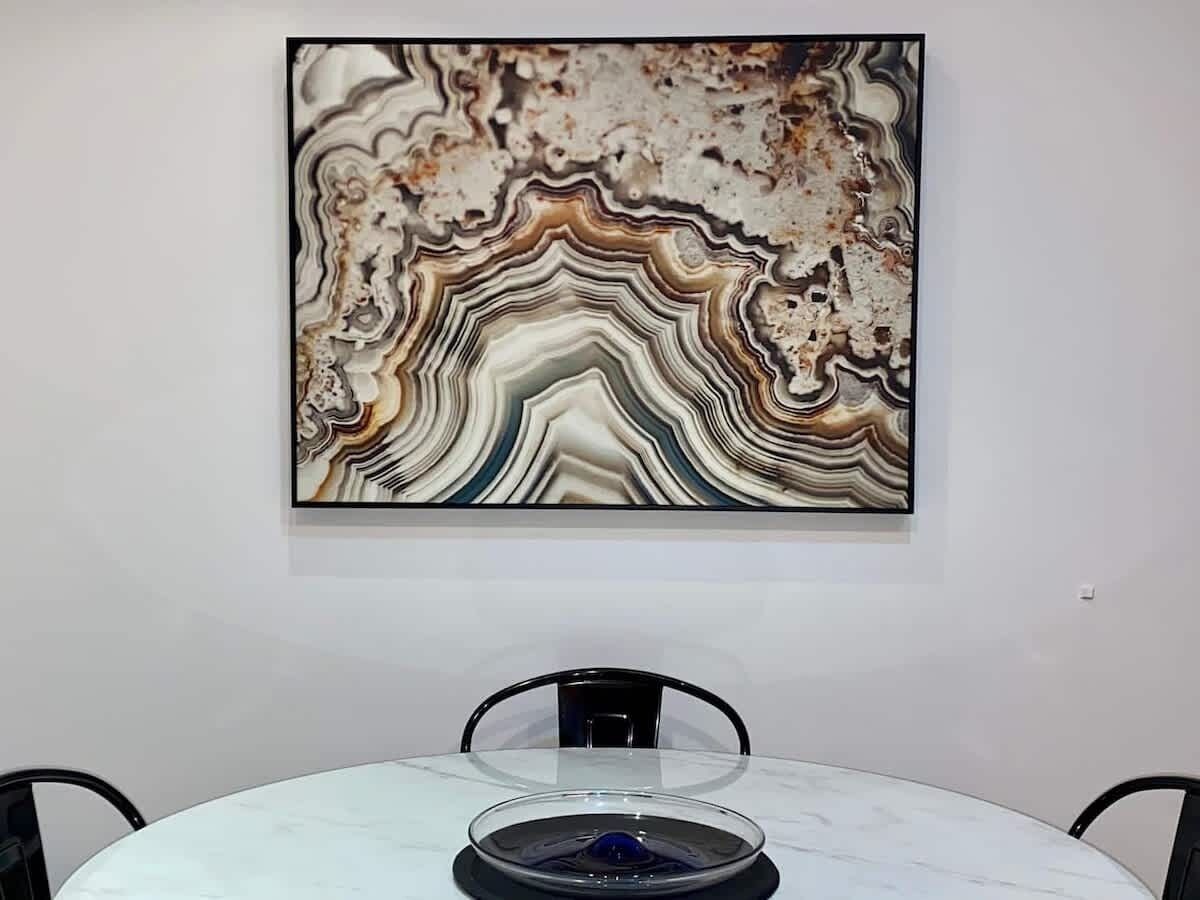 A room with a marble table and chairs, a framed abstract painting on the wall, and a small table with a blue object on it.